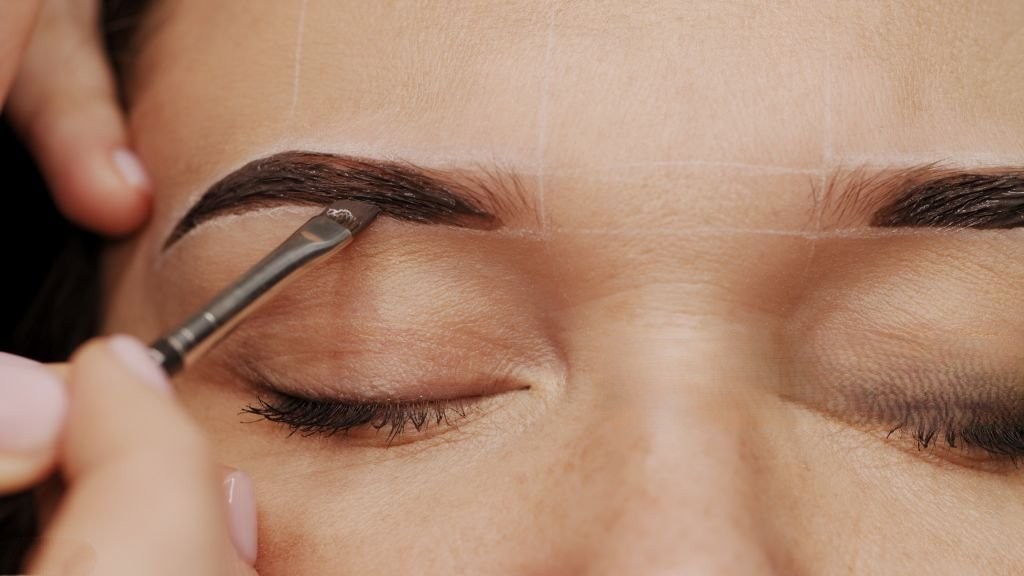 Woman is  on the Eyebrow coloring procedure. Professional lamination procedures of female eyebrows in beauty salon. Closeup view of client face and stylist's hands working.  Beauty care concept.